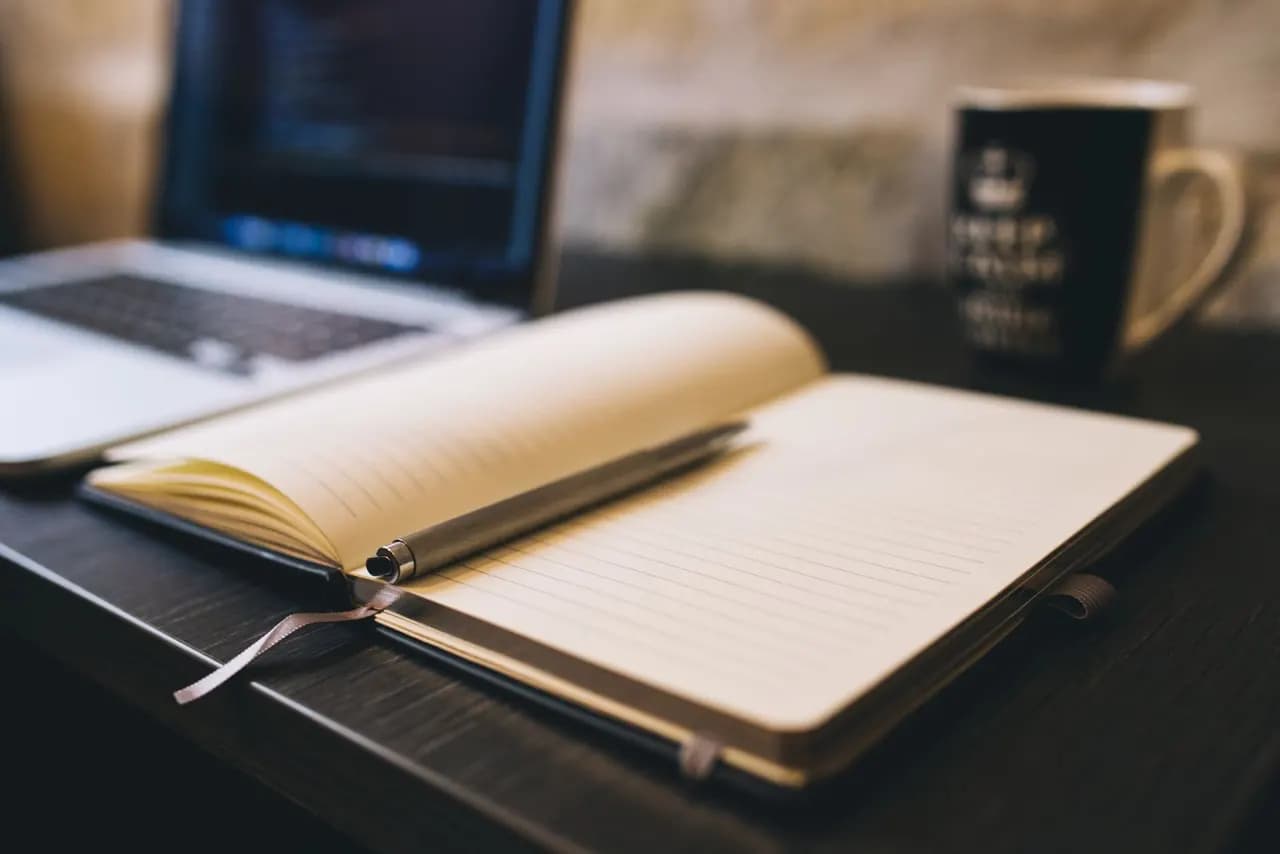 Dizüstü bilgisayarın yanında açık duran çizgili bir defter ve kalem